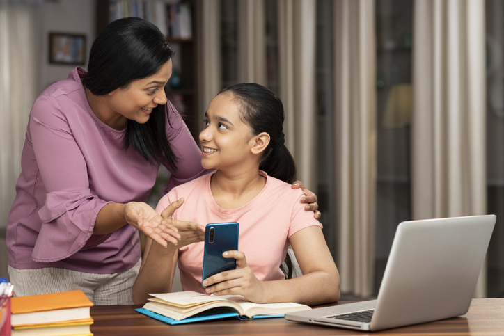 Teenage daughter showing mobile to her mom while studying at home